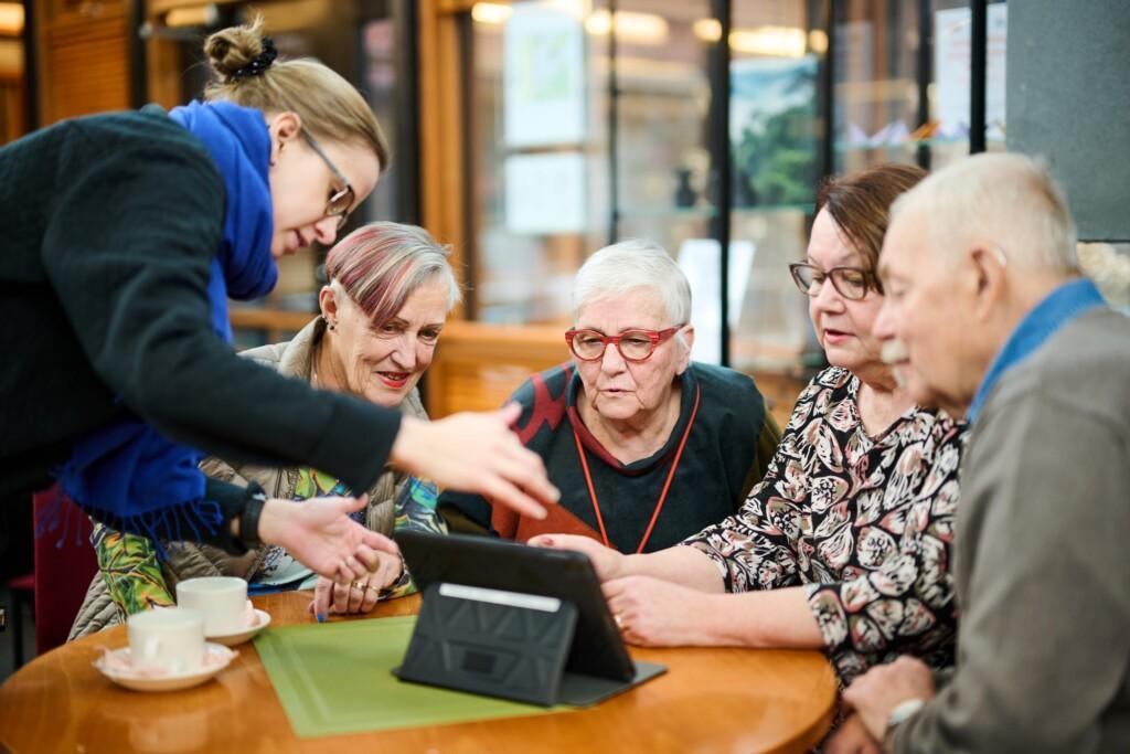 Kolme iäkästä naista ja yksi iäkäs mies tutkivat palvelutalon aulassa tablettia. Keski-ikäinen nainen ohjeistaa. Vanhustenviikko. Vanhustenpäivä Pukkila. 