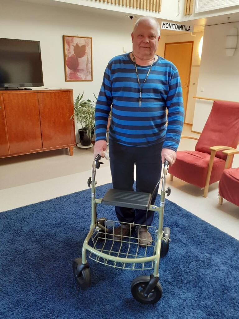 Iäkäs mies seisoo rollaattorin tukemana palvelutalon aulassa.