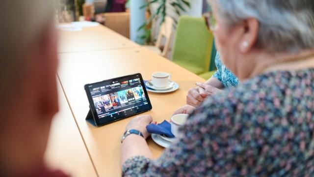 Ruokapöydällä tabletti, jota iäkäs nainen katselee kahvikupin äärellä