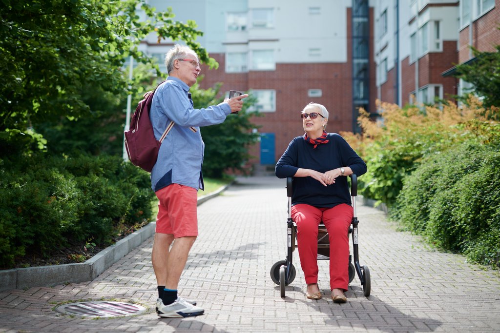 Mies ja nainen keskustelevat ulkona; nainen istuu pyörätuolissa ja mies seisoo vieressä pitäen juomaa kädessään.