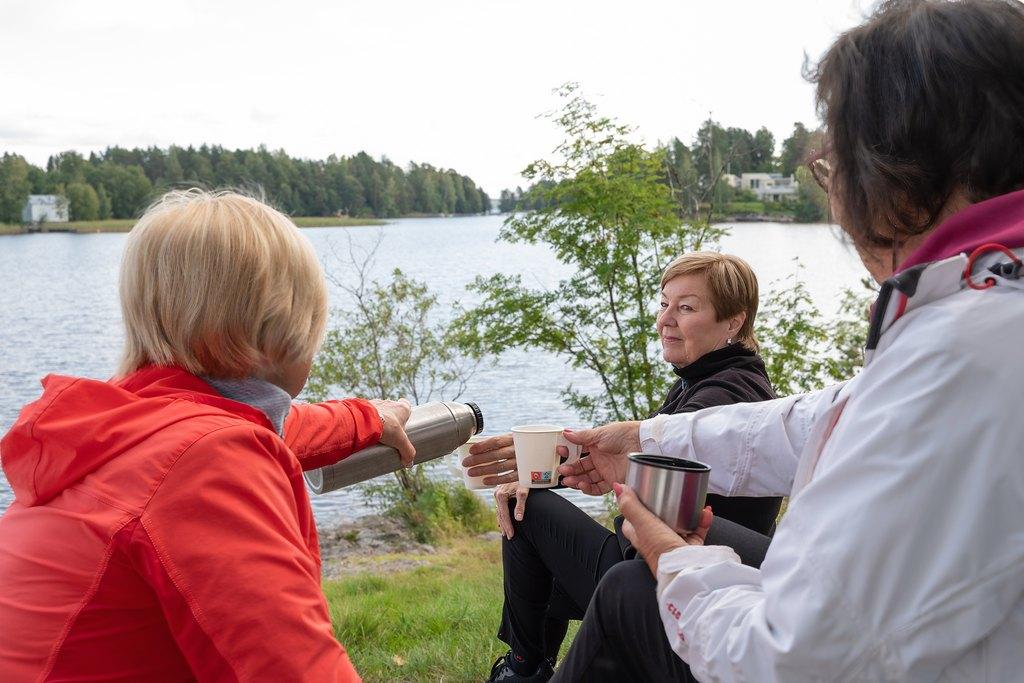 Kolme naista istuu järven rannalla ja juo kuumaa juomaa termospullosta mukiin kaadettuna.