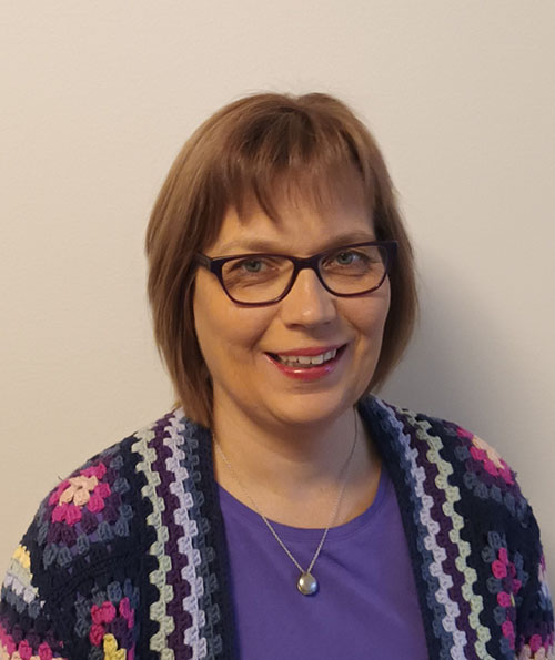 Hymyilevä silmälasipäinen nainen katsoo kameraan vaaleaa seinää vasten. Hänellä on violetti paita sekä värikäs, virkattu neuletakki, jossa on sinisen, violetin, pinkin ja valkoisen sävyjä. Kaulassa on hopeinen kaulakoru pyöreällä riipuksella.