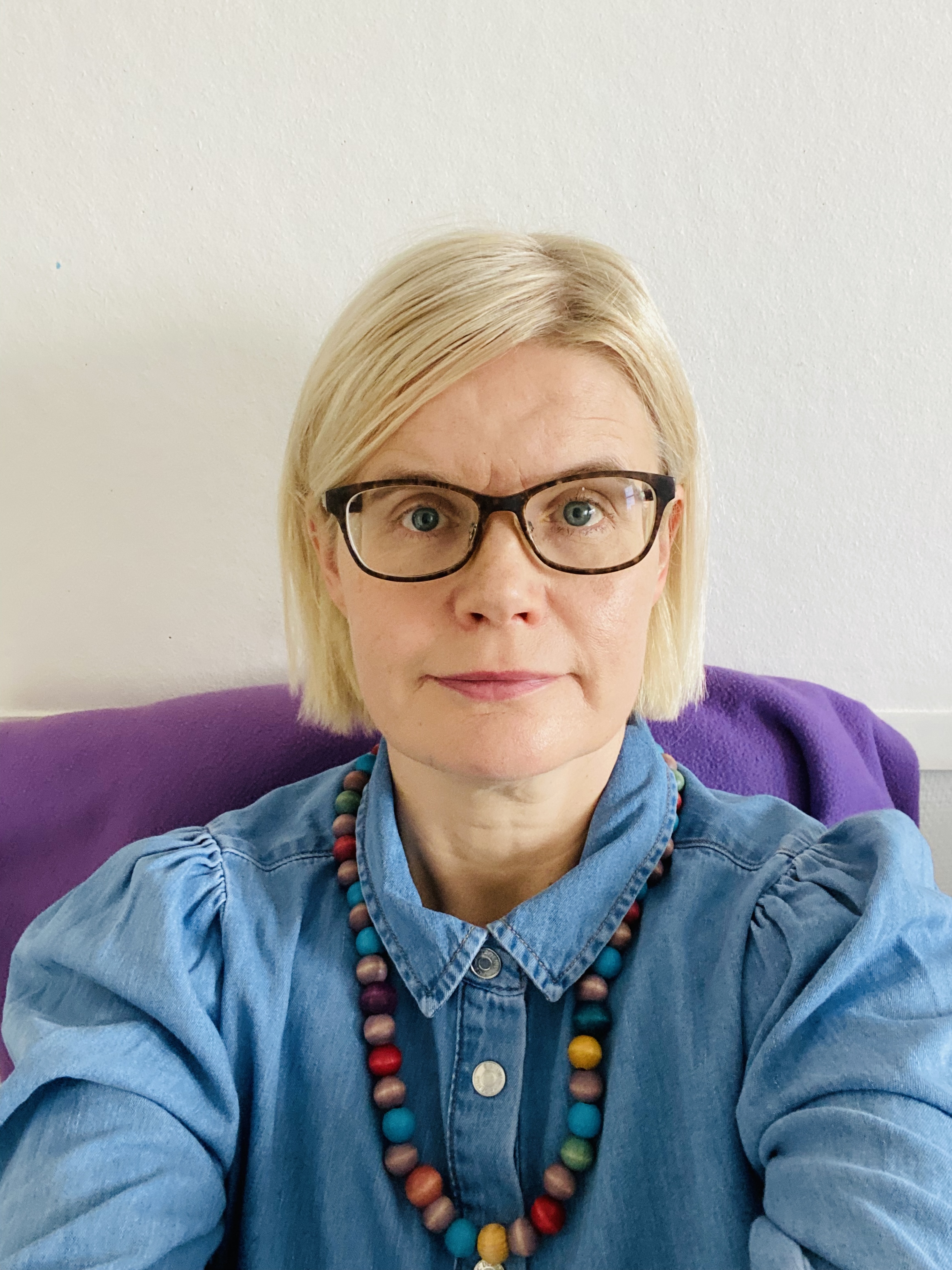 Silmälasipäinen vaaleahiuksinen nainen katsoo kameraan. Hänellä on sininen farkkupaita ja värikäs helmikaulakoru.