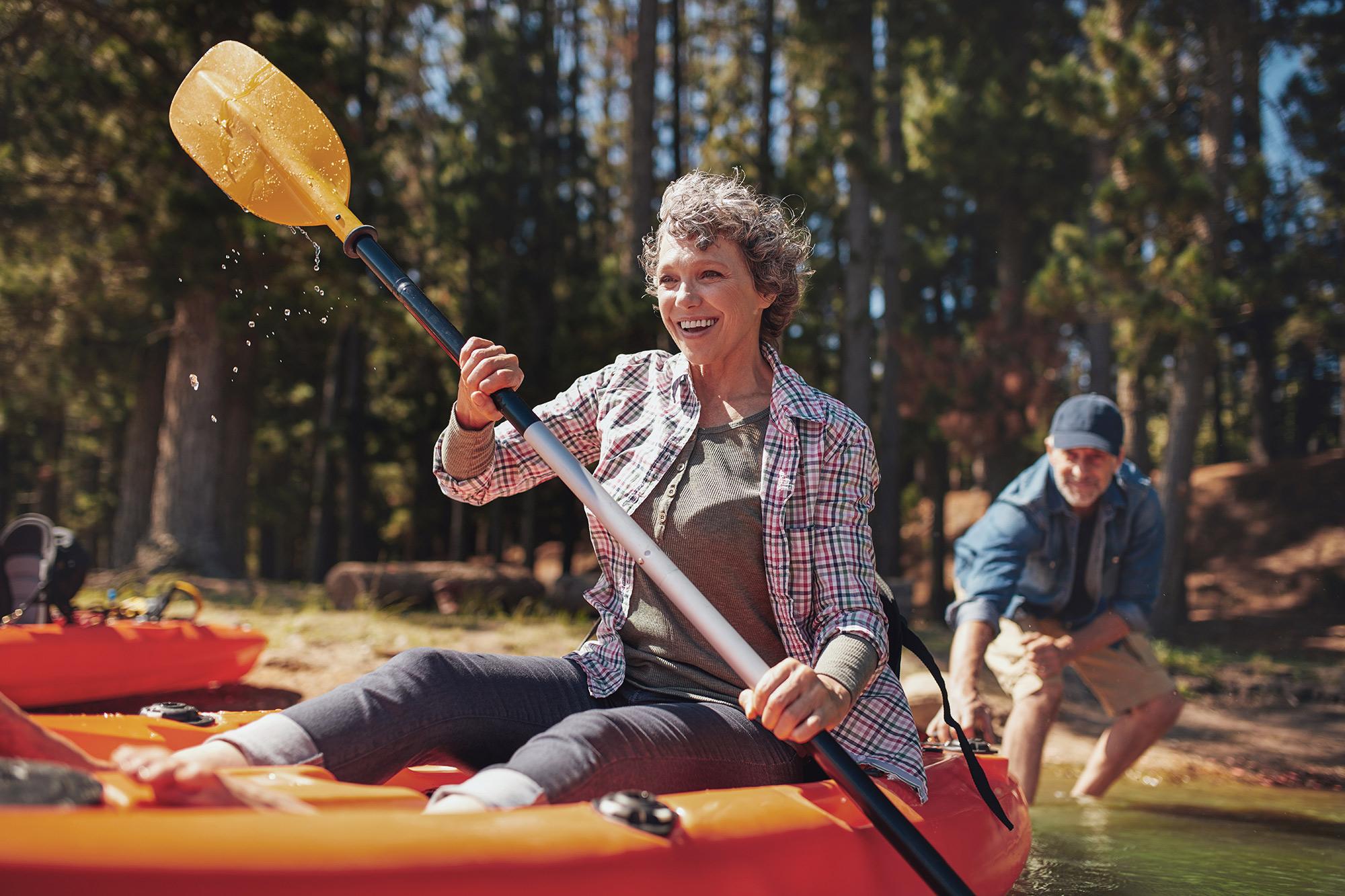 Keski-ikäinen nainen meloo kanootilla. Mies lykkää rannalta vauhtia.