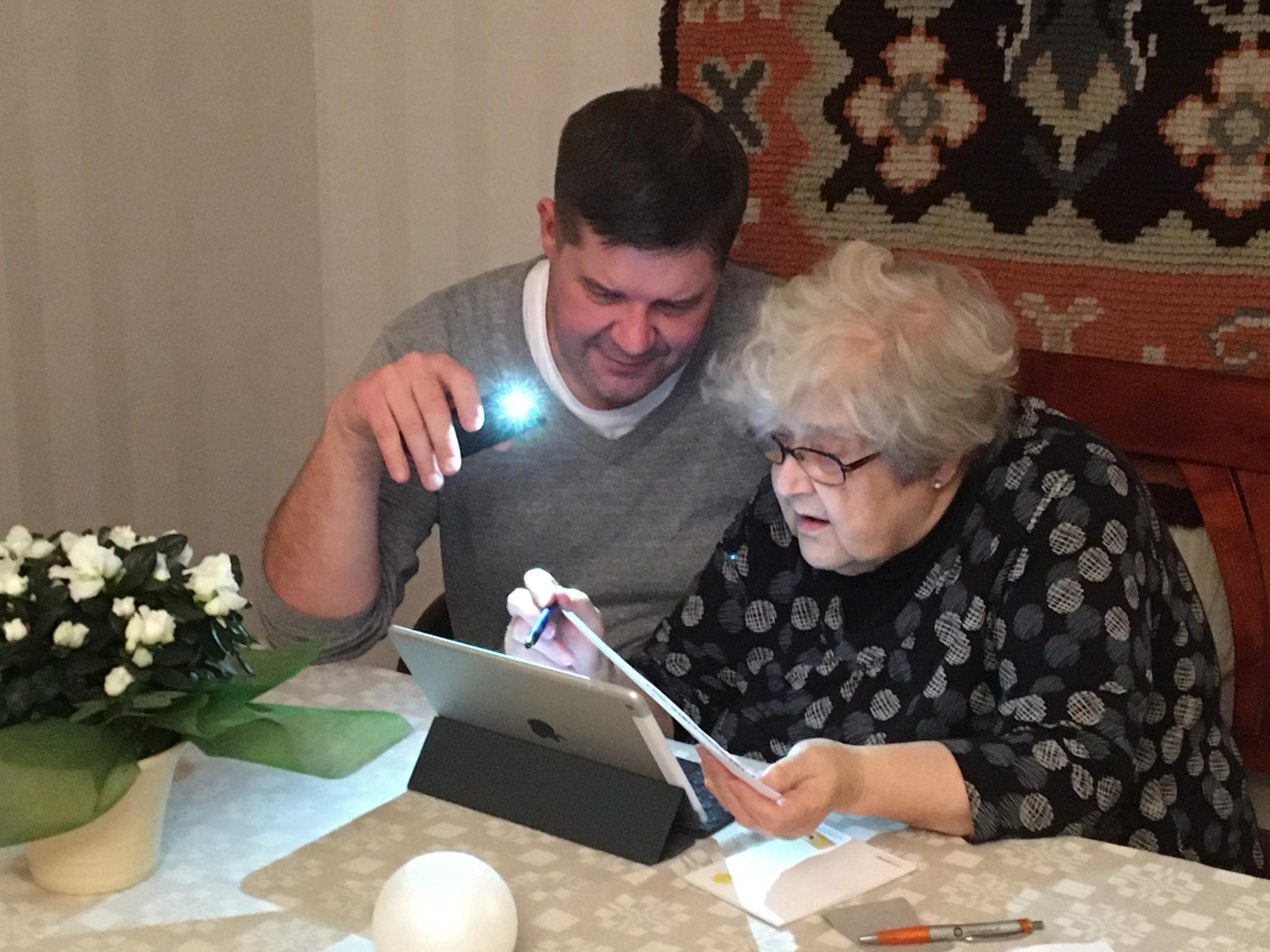 Keski-ikäinen mies opastaa vanhaa naista maksamaan laskuja tietokoneella.