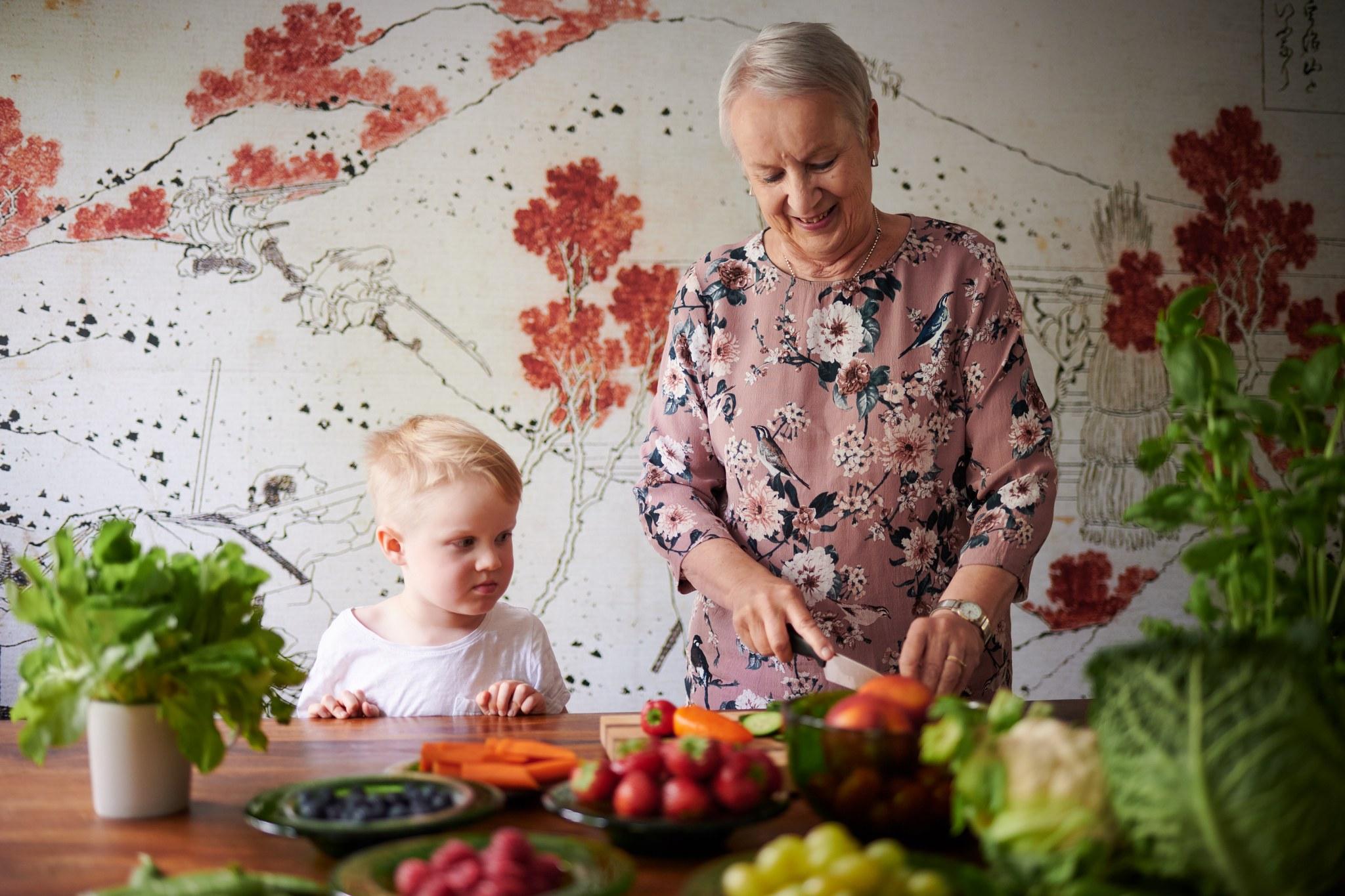 Isoäiti leikkaa pöydän ääressä vihanneksia ja pieni lapsi katsoo vierestä.