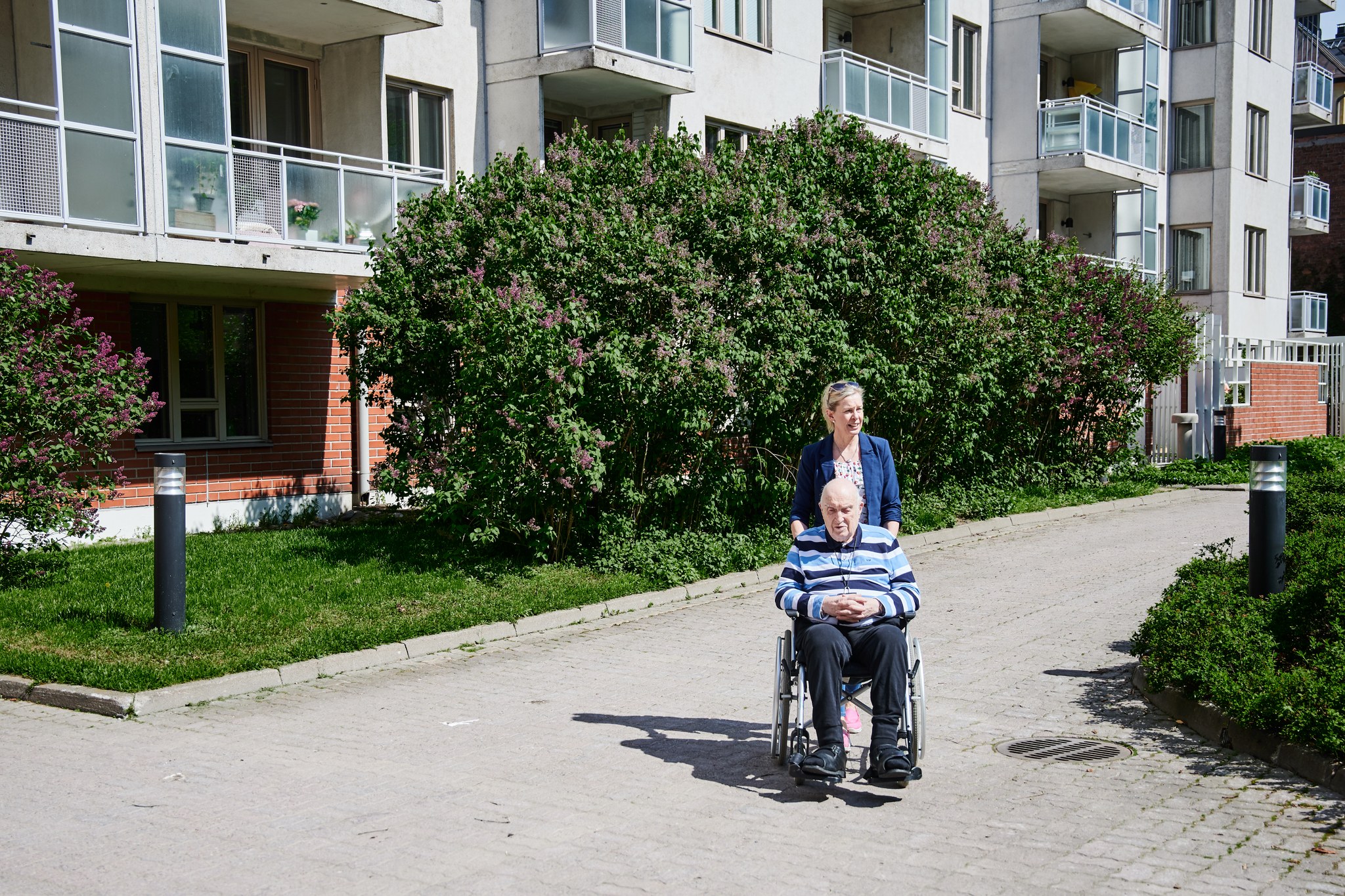 Keski-ikäinen nainen työntää iästä miestä pyörätuolissa kerrostaloalueen sisäpihalla. On kesä.