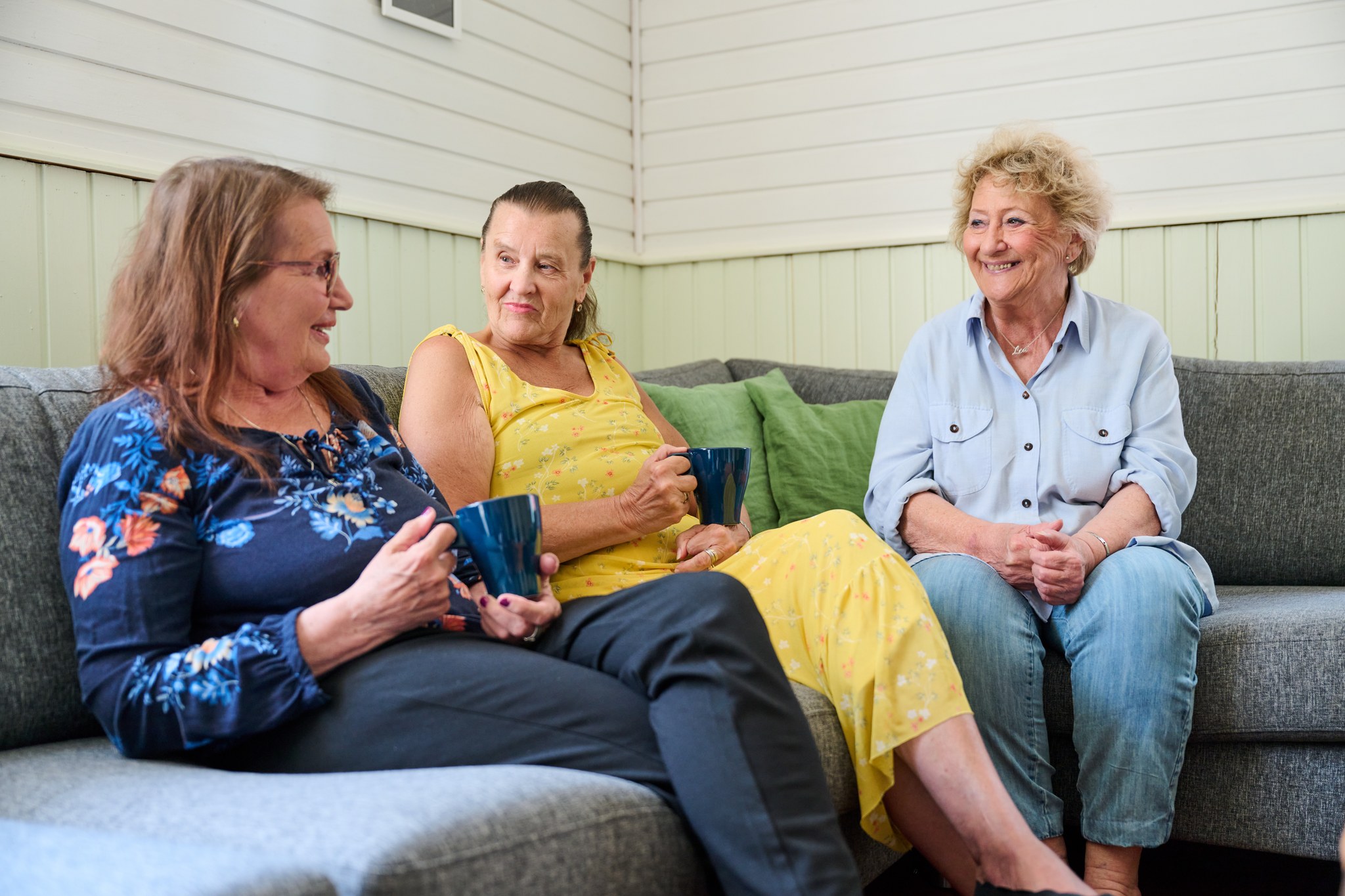 Kolme iäkästä naista istuu sohvalla juttelemassa. Kahvikupit kädessä.