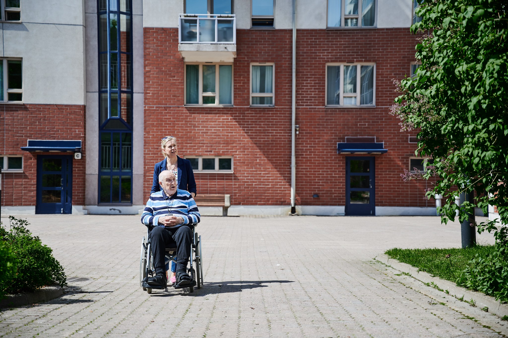 Nainen työntää iäkästä miestä pyörätuolissa kerrostalon pihalla.