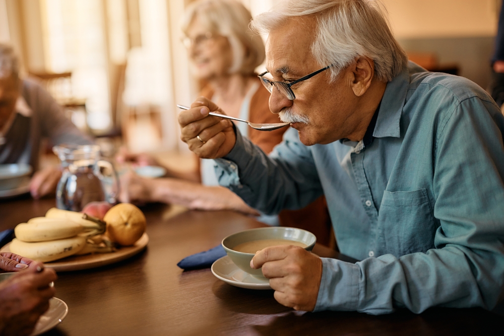 Iäkäs mies syömässä keittoa. Taustalla näkyy vanha nainen.