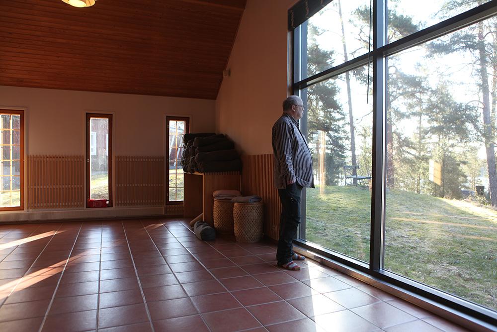 Iäkäs mies seisoo suuren ikkunan edessä ja katsoo metsämaisemaa.