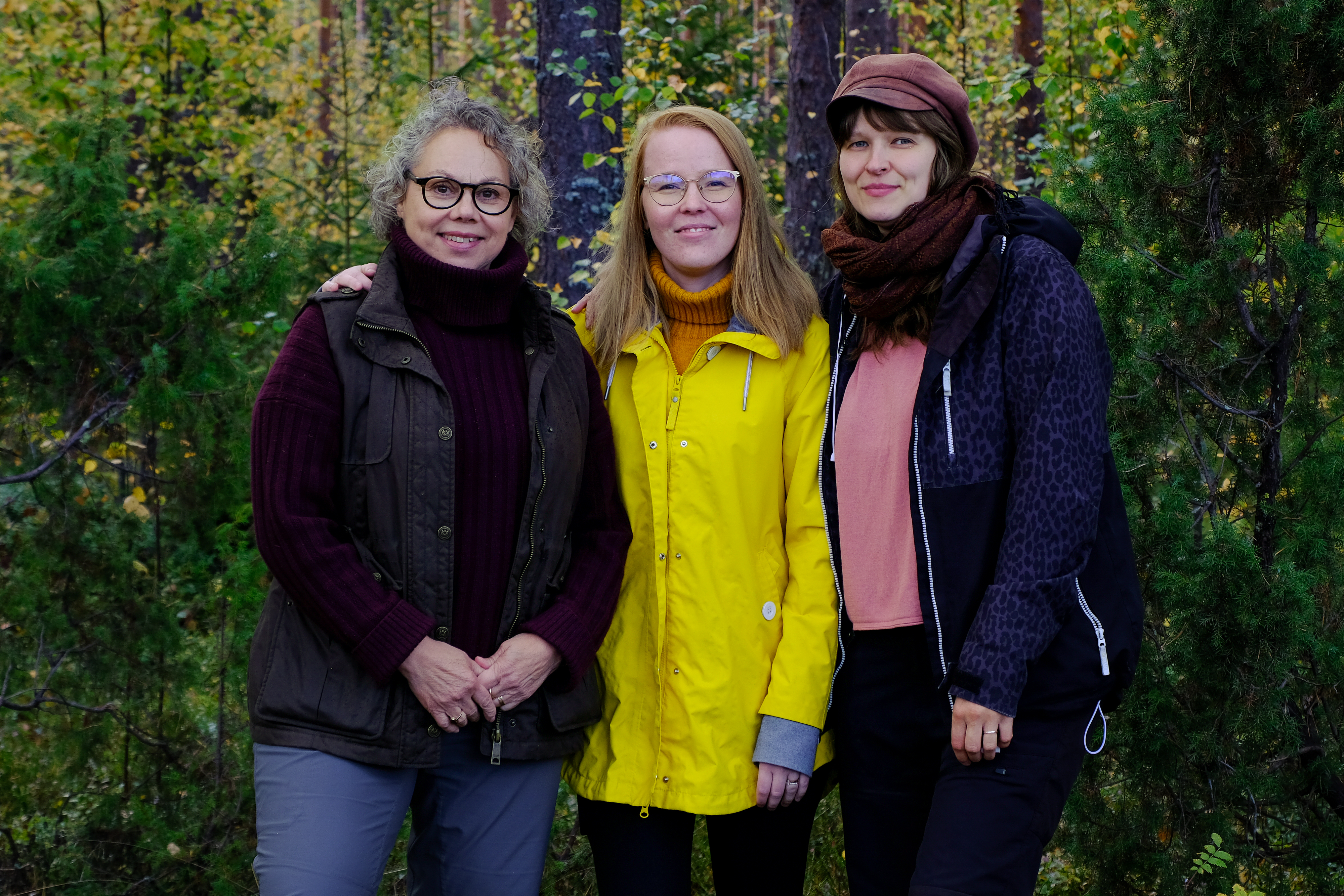 Kolme naista seisoo vierekkäin metsäisessä ympäristössä. He katsovat kameraan ja hymyilevät, keskellä oleva henkilö pukeutunut kirkkaankeltaiseen takkiin.