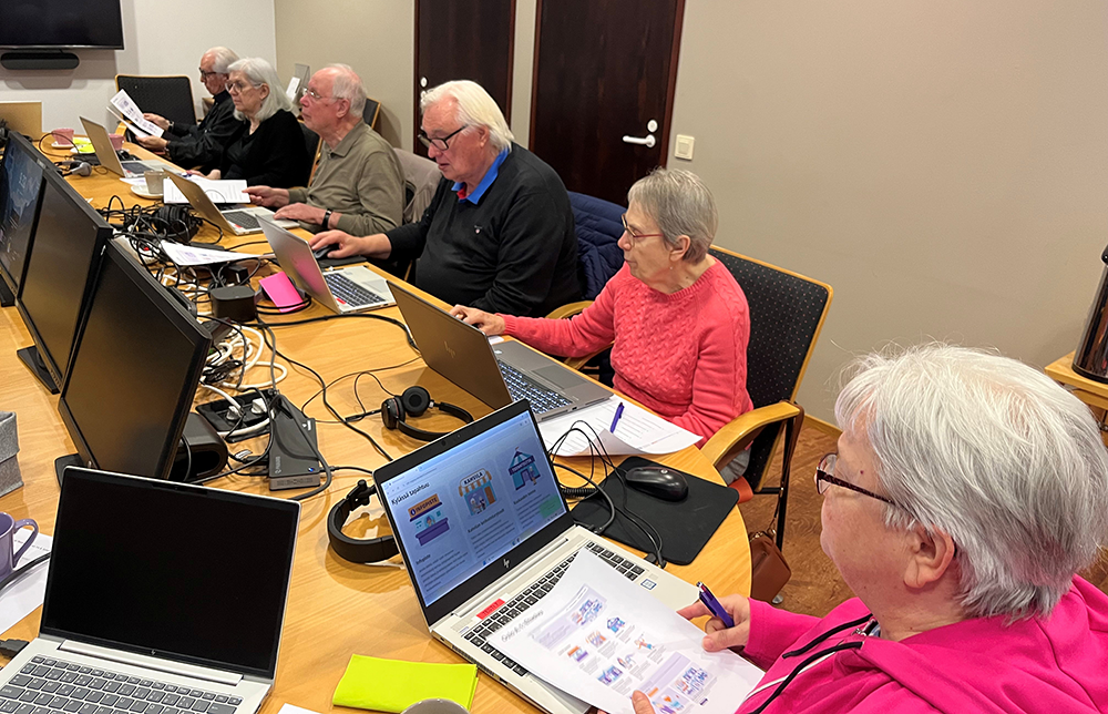 Ryhmä iäkkäitä ihmisiä istuu pöydän ääressä koulutustilassa ja käyttää kannettavia tietokoneita. He seuraavat opetusta, tekevät muistiinpanoja ja työskentelevät keskittyneesti. Pöydällä on tietokoneita, hiiriä, kuulokkeita ja papereita.