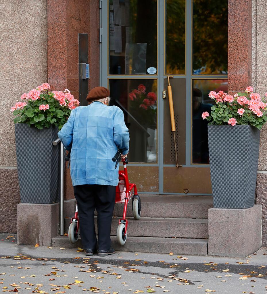 Vanhus menee rollaattorin kanssa talon ulkorappusia ylös ovelle. 