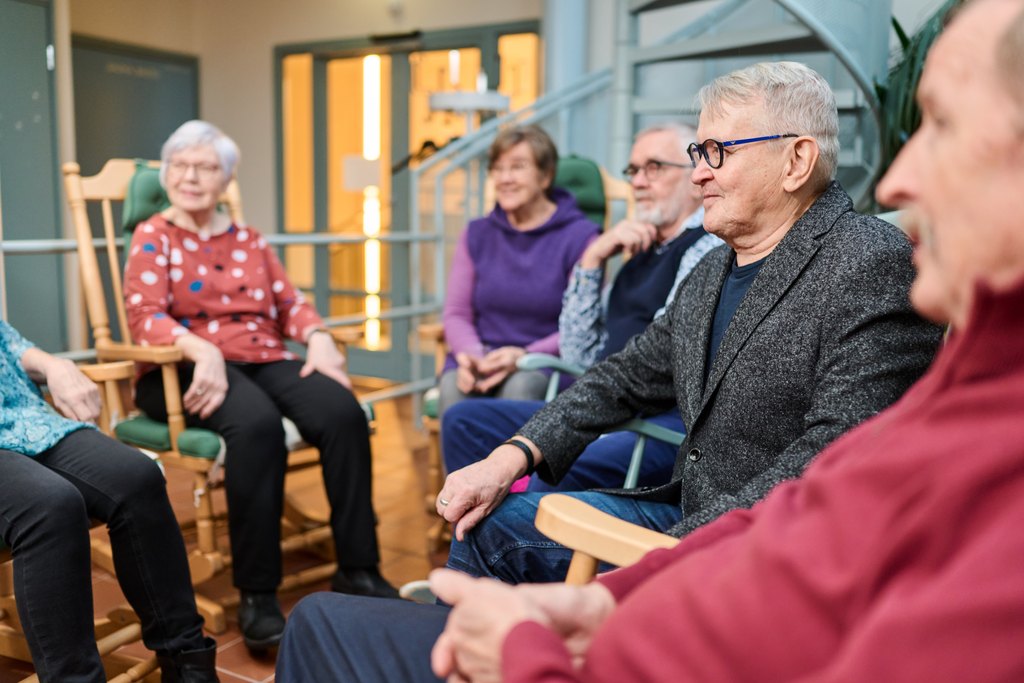 En grupp människor i olika åldrar sitter i en cirkel i en trivsam inomhusmiljö och samtalar. I bakgrunden syns en spiraltrappa och gröna dörrar. Flera deltagare sitter i gungstolar av trä och verkar koncentrerade och intresserade.