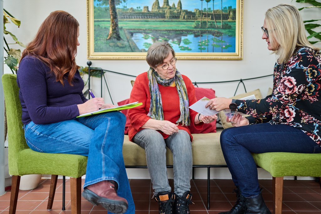 Kaksi naista haastattelee iäkästä henkilöä sohvalla.