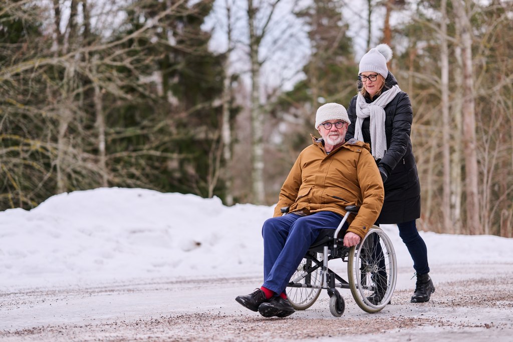 Talvinen ulkoilutilanne, jossa nainen työntää pyörätuolissa istuvaa iäkästä miestä lumisen tien reunassa. Molemmat ovat pukeutuneet lämpimiin vaatteisiin, ja he näyttävät nauttivan yhdessäolosta raikkaassa ulkoilmassa. Taustalla näkyy puita ja metsää.
