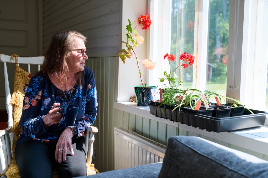 Iäkäs nainen istuu keinutuolissa ja katsoo ikkunasta ulos pitäen kädessään kahvikuppia. Ikkunalaudalla on kukkia ja taimilaatikoita, ja aurinko paistaa sisään huoneeseen, luoden lämpimän ja rauhallisen tunnelman
