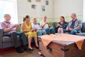 Kuusi henkilöä istuu yhdessä harmaalla sohvalla viihtyisässä huoneessa. He kaikki pitävät pelikortteja käsissään ja juttelevat keskenään hyväntuulisesti. Edessä on pöytä, jolla on oranssi liina, vesikannu ja sinisiä mukeja. Taustalla valkoisia paneloituja seiniä, ikkuna ja valokuvia seinällä.