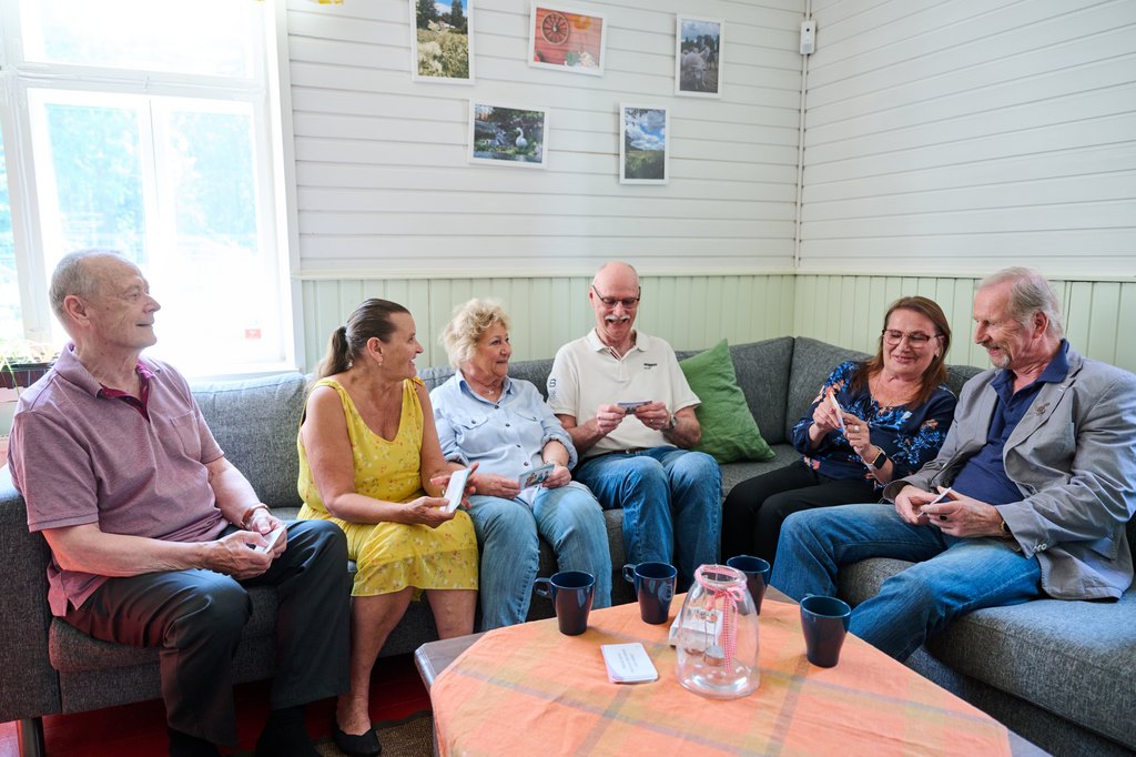 Kuusi henkilöä istuu yhdessä harmaalla sohvalla viihtyisässä huoneessa. He kaikki pitävät pelikortteja käsissään ja juttelevat keskenään hyväntuulisesti. Edessä on pöytä, jolla on oranssi liina, vesikannu ja sinisiä mukeja. Taustalla valkoisia paneloituja seiniä, ikkuna ja valokuvia seinällä.