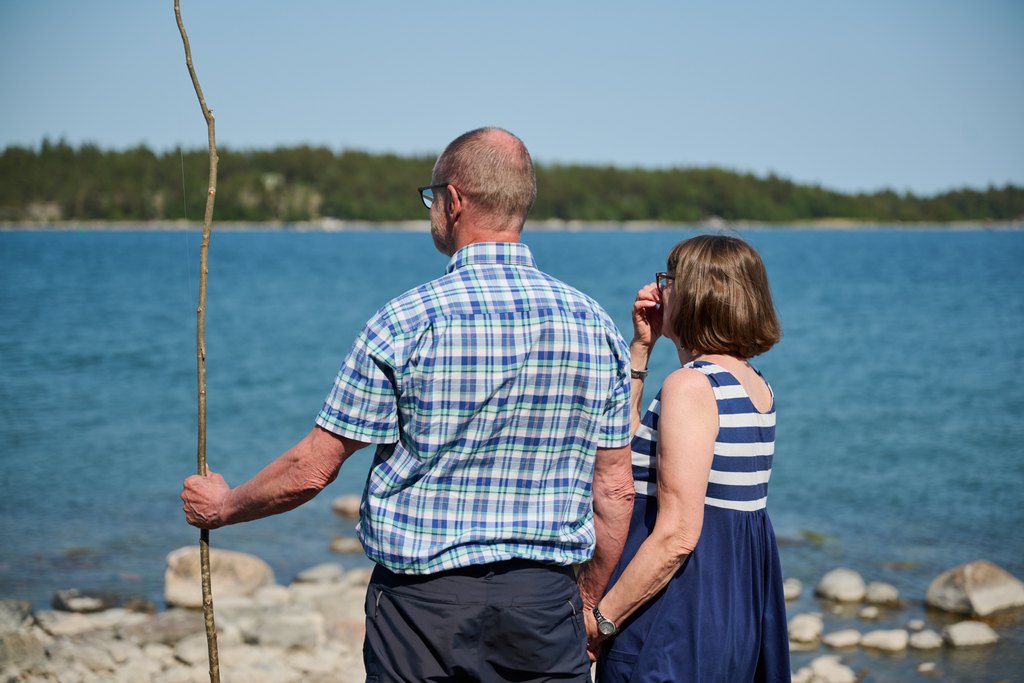 Ett äldre par står på klipporna vid havet och ser ut över vattnet. Bredvid dem står en picknickkorg och en blå tröja. Solen skiner från en klarblå himmel och i bakgrunden syns små öar.