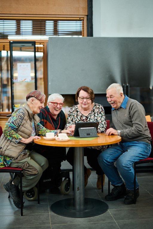 Äldre personer sitter i röda fåtöljer i en hemtrevlig miljö, läser en bok och använder en surfplatta, på bordet finns kaffekoppar och en blomma vid fönstret.