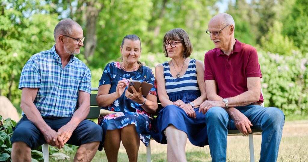Ryhmä iäkkäitä henkilöitä istuu penkillä ulkona tuijottaen puhelinta.