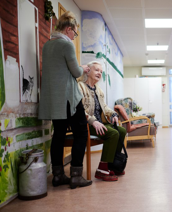 Hoivakodin käytävällä istuva iäkäs nainen saa apua toiselta henkilöltä, joka seisoo hänen vieressään. Taustalla seinään on maalattu värikäs seinämaalaus, jossa näkyy muun muassa kissa ja maisemaa. Käytävässä on myös nojatuoleja ja maitotonkka koristeena.