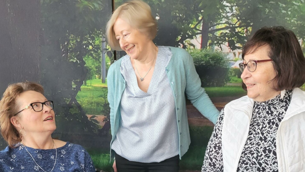 Kolme naista seisoo ja istuu yhdessä keskustellen ja hymyillen. Keskellä oleva nainen seisoo ja katsoo iloisena toisia, kun taas molemmat reunimmaiset naiset istuvat ja katsovat toisiaan ystävällisesti. Taustalla on seinä, jossa on valokuvamainen metsäinen maisema.