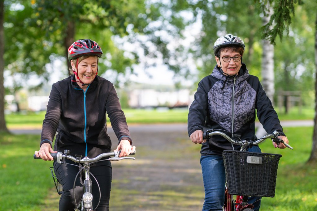 Två äldre kvinnor cyklar bredvid varandra i en parkmiljö, båda har cykelhjälmar och ett avslappnat, glatt uttryck.