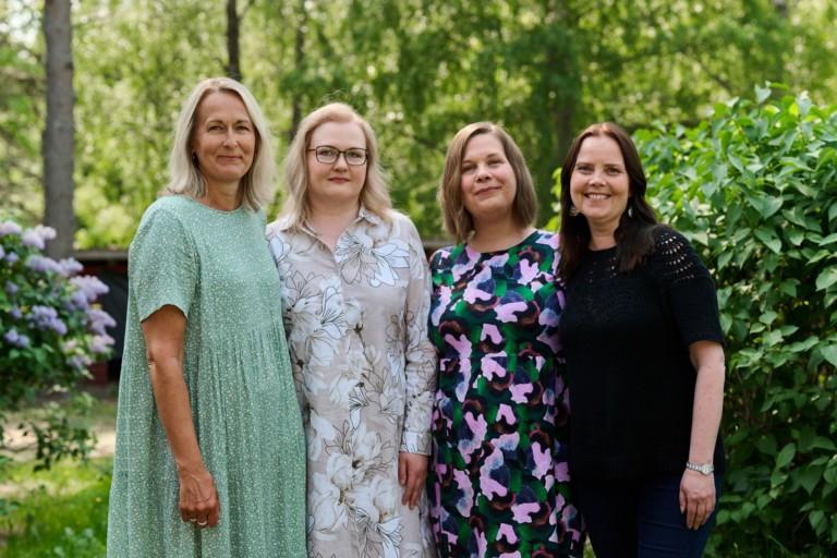 Neljä naista seisoo vierekkäin ulkona vehreässä ympäristössä, taustalla puita ja pensaita. Kaikki hymyilevät ja katsovat kameraan. He ovat pukeutuneet kesäisiin asuihin, kuten mekkoihin ja puseroon.