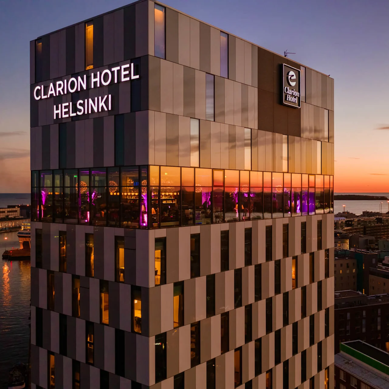 Clarion Hotel Helsinki -hotellin moderni tornirakennus iltavalaistuksessa, ikkunoista heijastuu auringonlasku ja kaupungin sekä meren maisema.