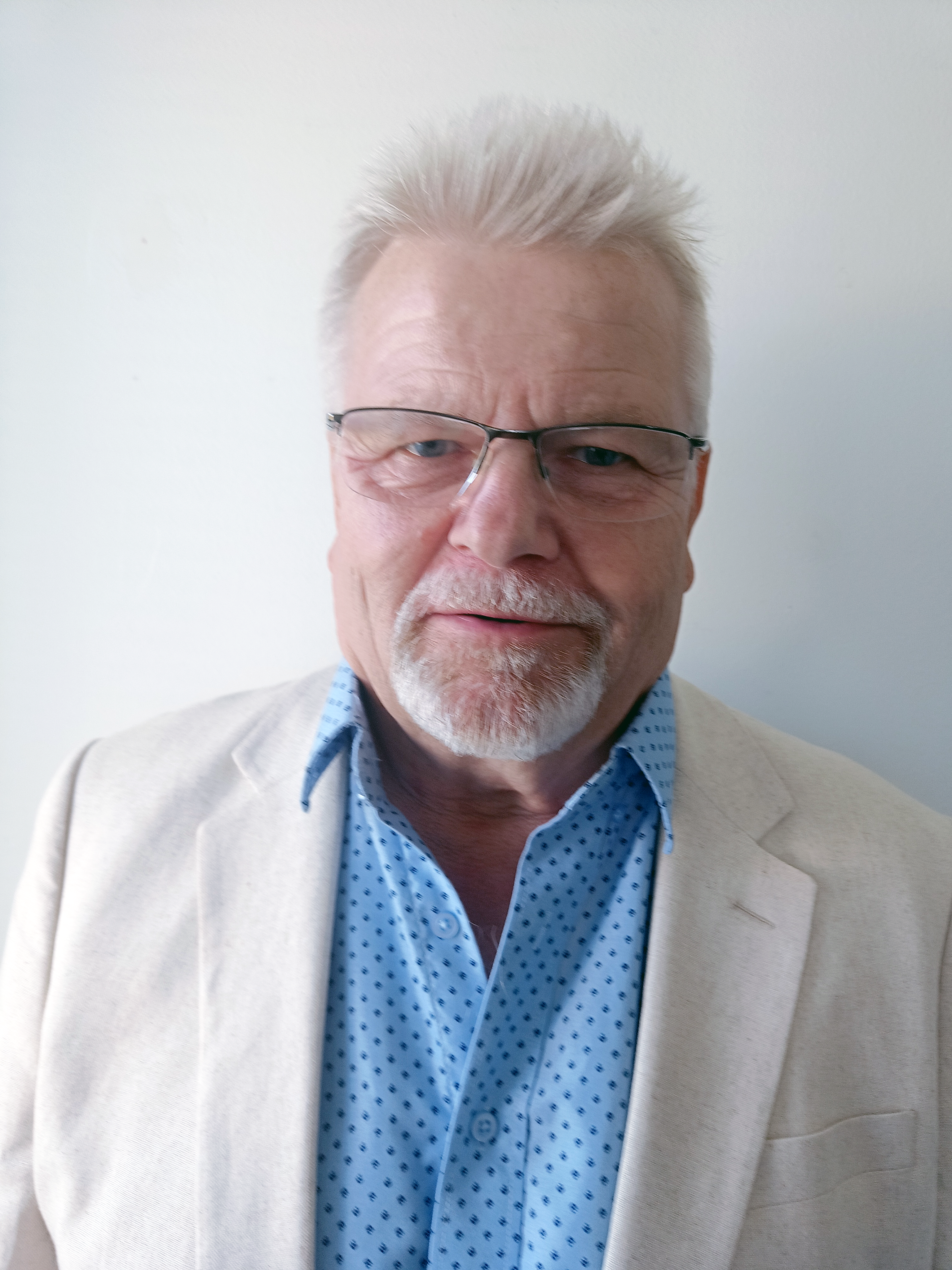 Vaaleaan pikkutakkiin ja siniseen kauluspaitaan pukeutunut harmaahiuksinen, silmälasipäinen mies katsoo kameraan vaaleaa seinää vasten.