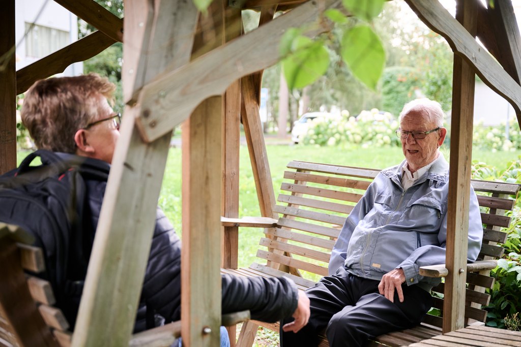 Kaksi miestä istuu puutarhakeinussa ulkona keskustelemassa. Vanhempi mies harmaassa takissa istuu keinun toisella puolella ja puhuu hymyillen. Nuorempi mies, selkä kameraan päin, kuuntelee tarkkaavaisesti. Taustalla näkyy vihreä piha ja puita.