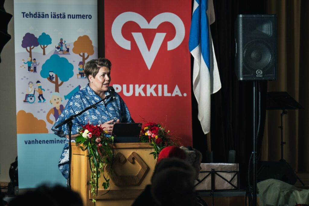 Nainen puhuu juhlayleisölle puhujanpöntöstä. Taustalla Pukkilan kunnan ja vanheneminen.fi:n visuaaliset elementit ja Suomen lippu.