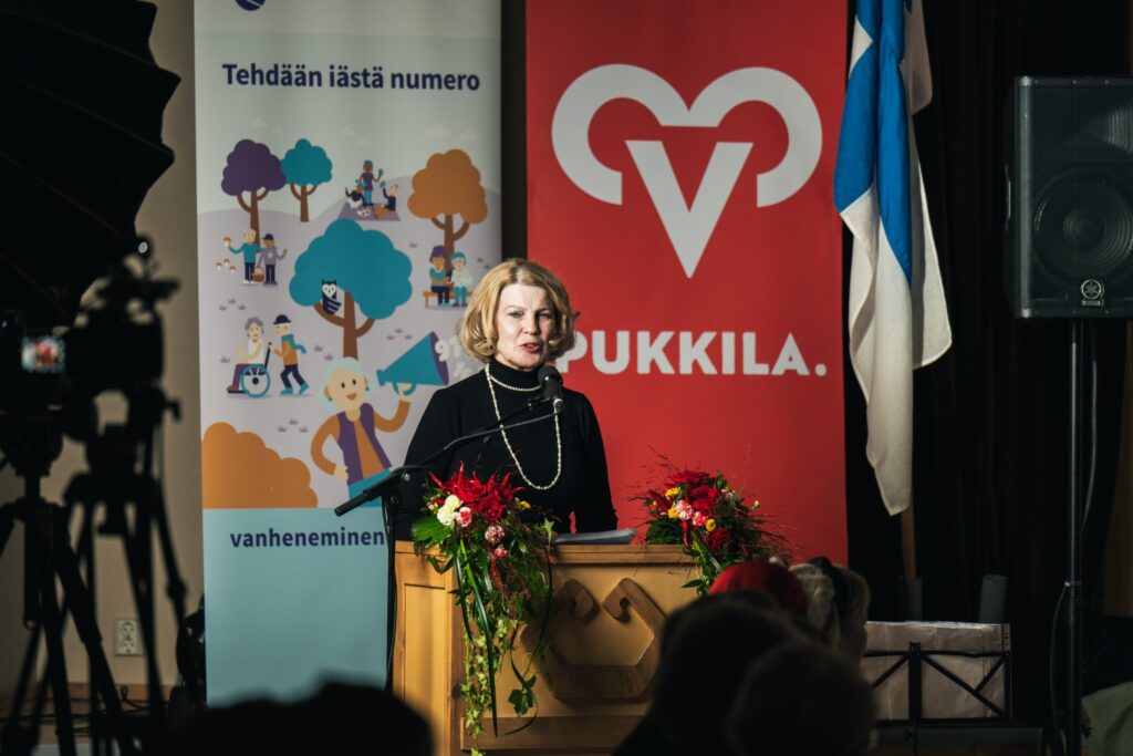 Nainen puhuu juhlayleisölle puhujanpöntöstä. Taustalla Pukkilan kunnan ja vanheneminen.fi:n visuaaliset elementit ja Suomen lippu.
