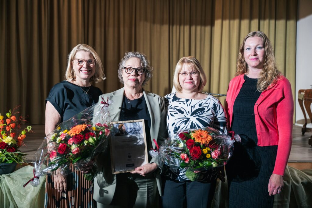 Neljä naista seisoo rivissä ja pitää käsissään kukkakimppuja ja kunniakirjaa. Heistä kolme on saanut huomionosoituksen. Taustalla ruskea verho.