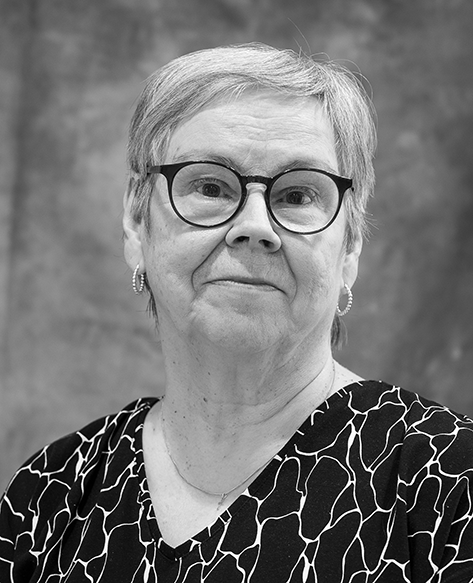 Mustavalkoinen muotokuva iäkkäästä naisesta, joka katsoo suoraan kameraan. Hänellä on silmälasit, korvakorut ja kuvioitu pusero. Taustana on neutraali studiossa otettu tausta.