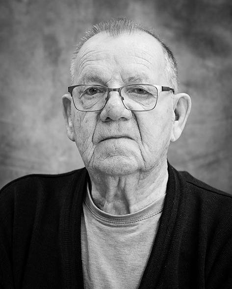 Mustavalkoinen muotokuva iäkkäästä miehestä, joka katsoo suoraan kameraan vakavailmeisenä. Hänellä on silmälasit, lyhyt tukka ja yllään kevyt paita sekä neuletakki. Taustana on pehmeäsävyinen kangas.