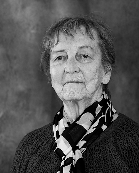 Mustavalkoinen muotokuva iäkkäästä naisesta, joka katsoo suoraan kameraan rauhallisella ilmeellä. Hänellä on lyhyt tukka, neulepusero ja kaulassaan kuviollinen huivi. Taustalla on tasainen, pehmeäsävyinen kangas.