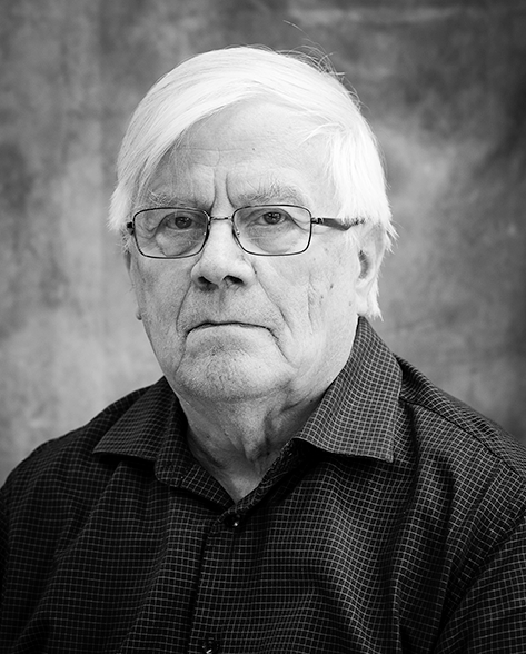 Mustavalkoinen muotokuva iäkkäästä miehestä, joka katsoo suoraan kameraan vakavasti. Hänellä on suorat, vaaleat hiukset, silmälasit ja ruudullinen kauluspaita. Taustalla on pehmeäsävyinen kangas.