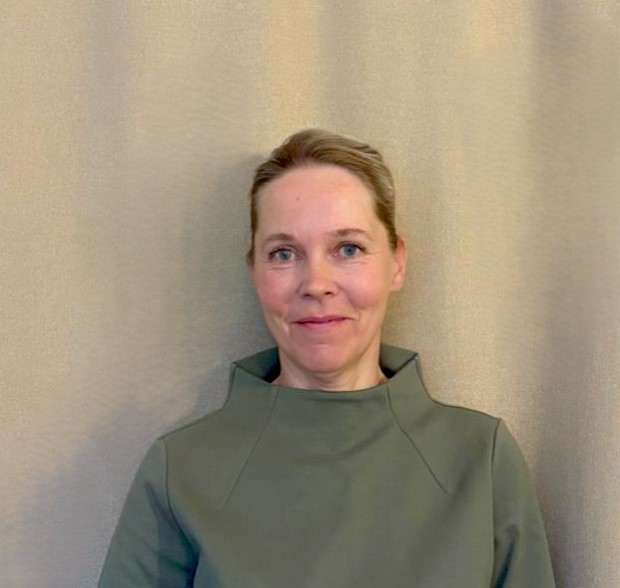 Henkilö seisoo tumman taustan edessä, katsoo kameraan sivuttain ja hymyilee kevyesti. Hänellä on tummansininen pusero ja punertavat kiharat hiukset.