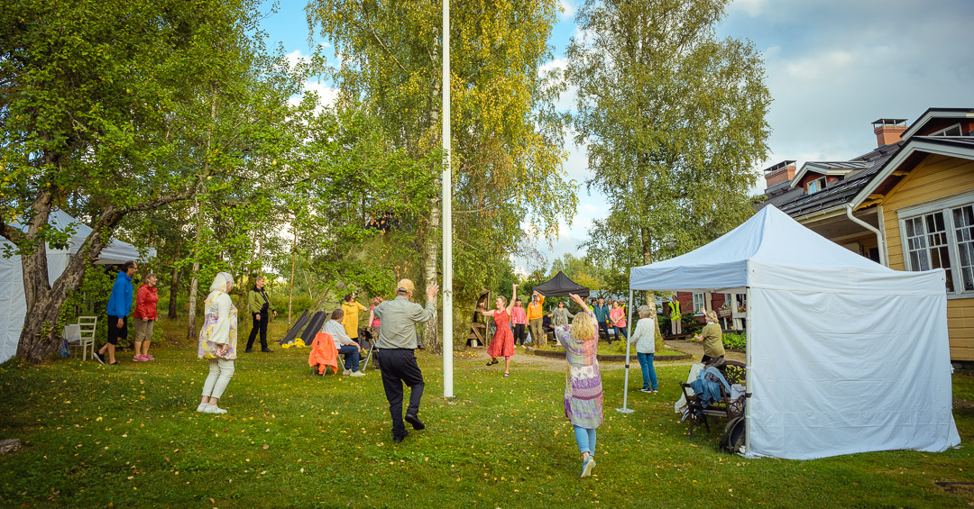 Puutarhatanssit ulkona vihreässä pihapiirissä. Iäkkäitä ja keski-ikäisiä ihmisiä osallistuu yhteiseen tanssituokioon lipputangon ympärillä. Osa katsoo tanssia, osa tanssii iloisesti musiikin tahdissa. Pihalla on pystytetty valkoisia ja mustia katoksia, ja taustalla näkyy vanhoja puutaloja. Tunnelma on yhteisöllinen ja hyväntuulinen.