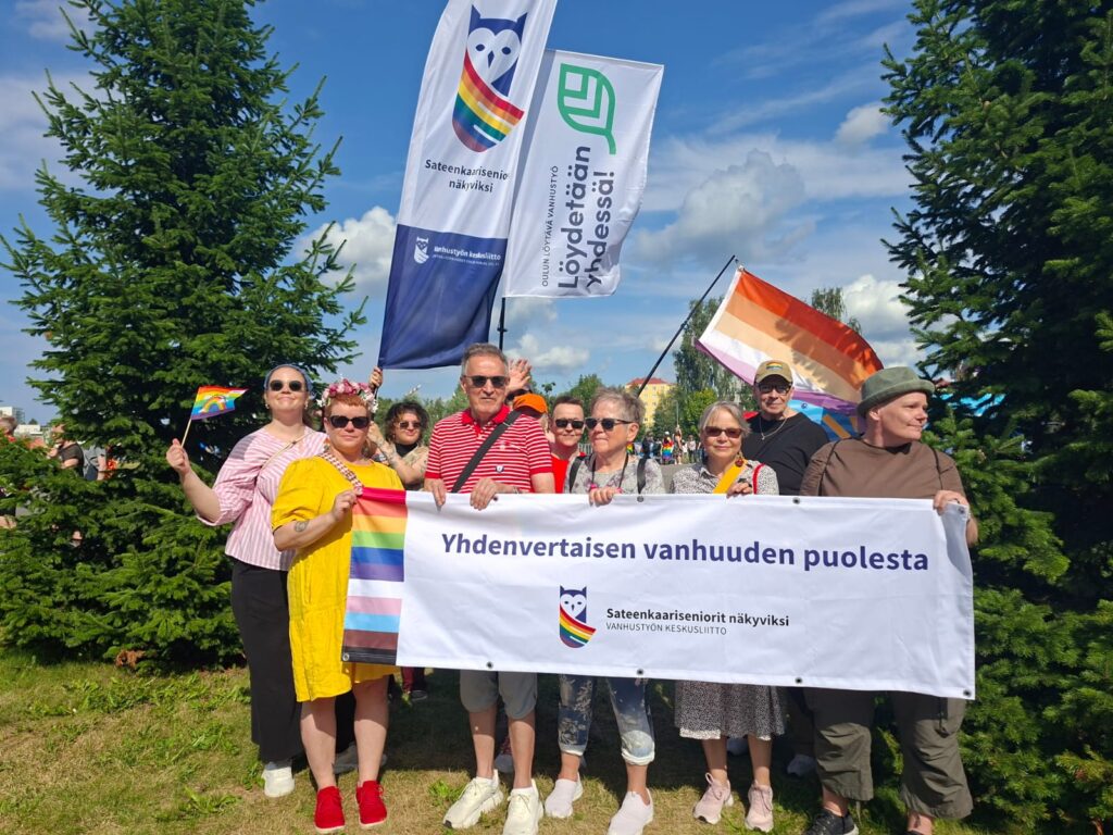 Joukko iloisia ihmisiä kesällä Sateenkaariseniorit näkyviksi -lipun ja banderollin kanssa.