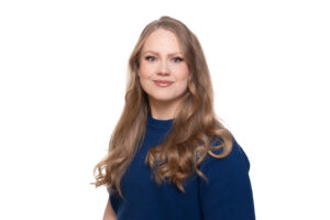 Värillinen muotokuva nuoresta aikuisesta, jolla on pitkät vaaleat hiukset ja sininen pusero. Hän katsoo kameraan ja hymyilee hillitysti valkoista taustaa vasten.