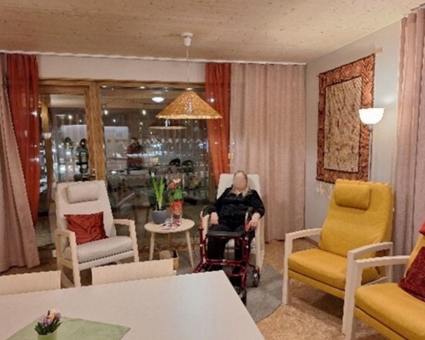 Kodikas olohuonemainen tila, jossa on nojatuoleja, sohvapöytiä ja sisustuselementtejä. Yksi ihminen istuu rollaattorin kanssa tuolilla. Huoneessa on suuret ikkunat verhoineen, koristeellinen seinätekstiili ja pöydällä kukkia.