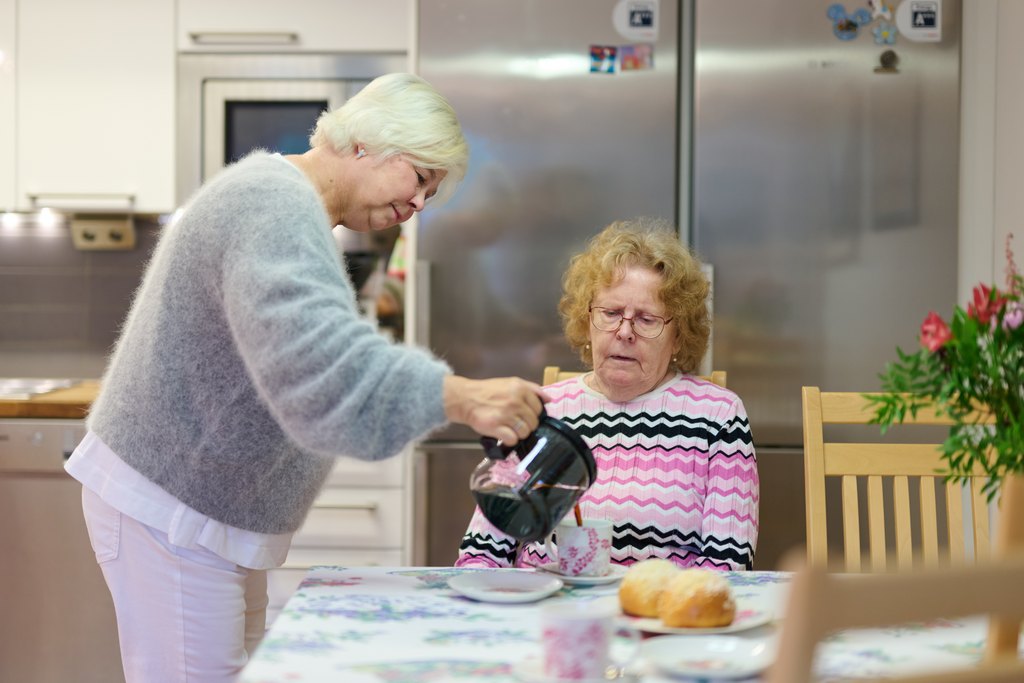 Kaksi iäkästä naista keittiössä. Toinen istuu pöydän ääressä kukkaliinan ja leivonnaisten äärellä, toinen kaataa kahvia kuppiin. Taustalla näkyy moderni keittiö ja jääkaappi.