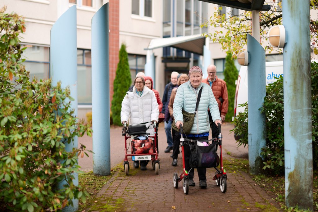 Ryhmä iäkkäitä ihmisiä kävelee ulkona jalkakäytävällä syksyisessä säässä. Etualalla kaksi naista työntää rollaattoria, toinen niistä punaisella rollaattorilla, jossa on nimikyltti ”MUMMI”. Taustalla näkyy useita muita kävelijöitä ja rakennuksen sisäänkäynti. Molemmin puolin on pensaita ja vaaleansiniset pylväät kehystämässä kulkureittiä.