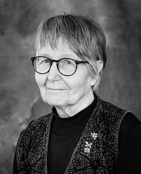 Mustavalkoinen muotokuva iäkkäästä naisesta, jolla on lyhyet, siististi leikatut hiukset ja pyöreät silmälasit. Hän katsoo kameraan lempeästi hymyillen ja käyttää tummaa puseroa sekä kuviollista liiviä. Tausta on pehmeä ja neutraali.