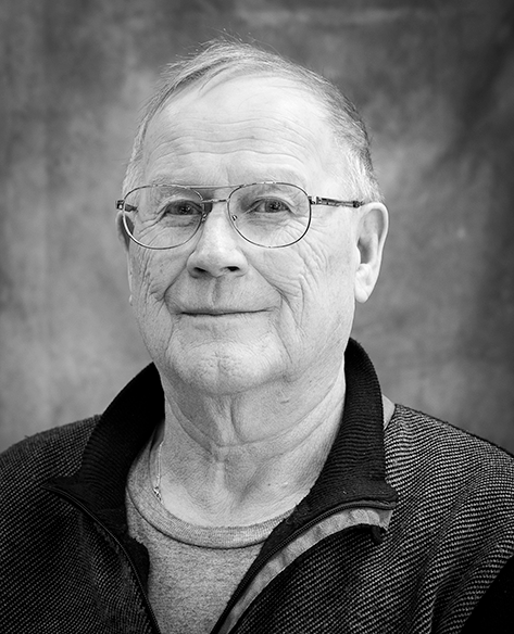 Mustavalkoinen muotokuva iäkkäästä miehestä, joka katsoo kameraan rauhallinen ilme kasvoillaan. Hänellä on puolireunaiset silmälasit ja yllään neulepaita, jonka alla on t-paita. Taustana on tasainen, utuisen pehmeä pinta.